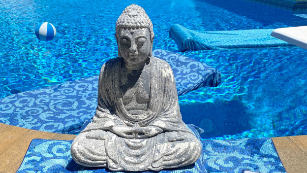 A Buddha statue sits on a Backyard Accents towel in front of a pool with a float and ball.