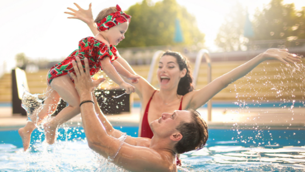 A man lifts a baby girl over his head in a pool as a woman cheers them on.