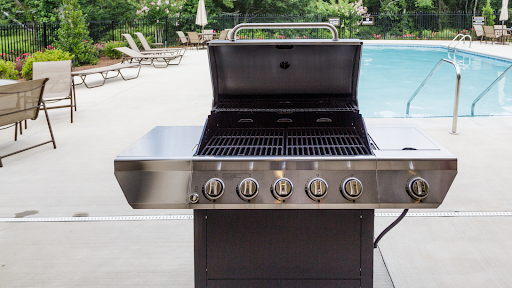 A silver and black grill is open and sits in front of a large pool.